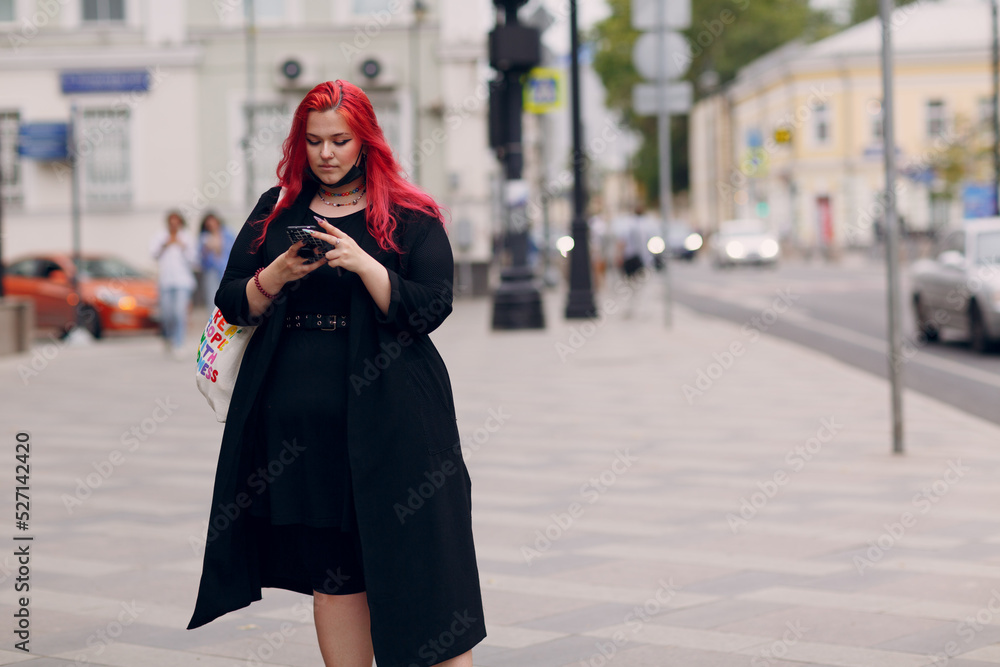 Fototapeta premium European plus size woman walking outdoor street city with mobile cellular phone. Young red pink haired body positive girl using cellphone smartphone