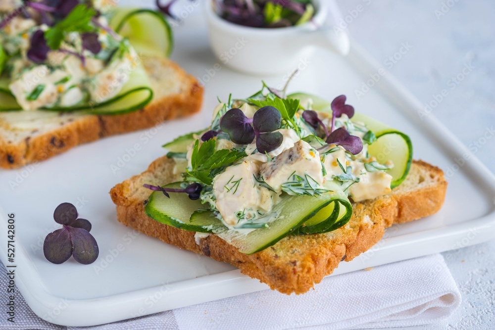 Open Danish smorrebrod sandwiches with chicken salad, fresh cucumber and herbs on a white ceramic board against a light concrete background. Sandwich recipes.