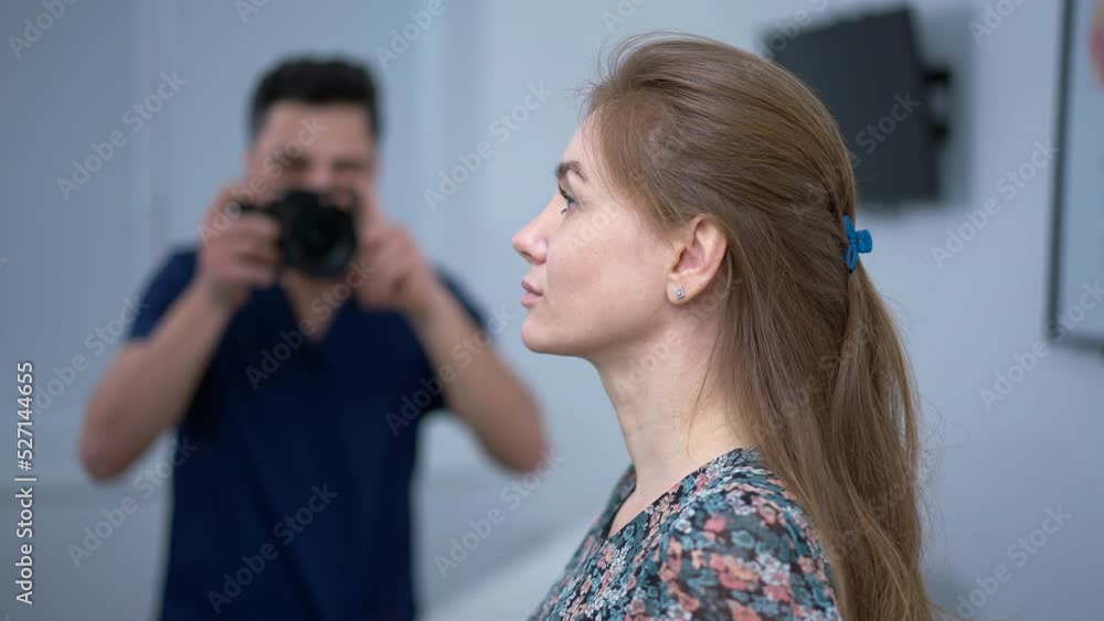 Side view slim adult Caucasian woman posing for before and after photo with blurred surgeon photographing with camera at background. Middle Eastern man taking photo of patient before plastic surgery Side view slim adult Caucasian woman posing for before and after photo with blurred surgeon photographing with camera at background. Middle Eastern man taking photo of patient before plastic surgery