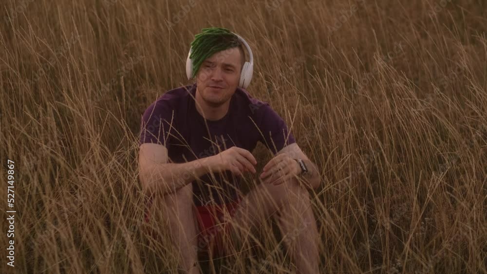 Young handsome man with green dreadlocks, wireless headphones listening to music in nature, sitting in dry grass.