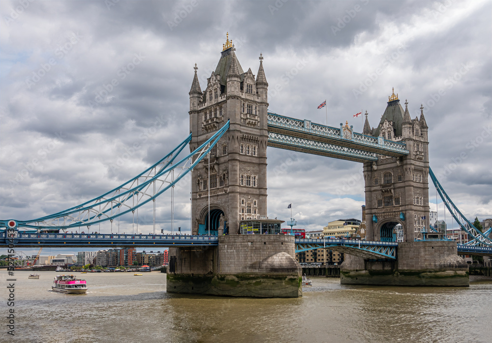 Fototapeta premium London, England, UK - July 6, 2022: Tower of London quay. Pink ferry sails in brown Thames water under north span of Tower bridge. Thick cloudscape above. 
