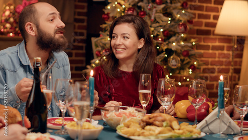 Festive married couple enjoying chatting with relatives while sitting at Christmas dinner table. Happy family celebrating winter feast with home cooked food and sparkling wine. Handheld shot