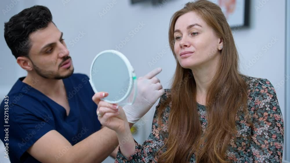 Middle Eastern man talking pointing at face of Caucasian woman looking at hand mirror. Expert plastic surgeon consulting patient on facelift procedure Middle Eastern man talking pointing at face of Caucasian woman looking at hand mirror. Expert plastic surgeon consulting patient on facelift procedure