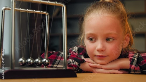 Concentrated child girl female little patient blonde cute beautiful small lady preschooler sitting on psychotherapy session indoors looking at swinging metal steel balls law kinetic energy hypnotized