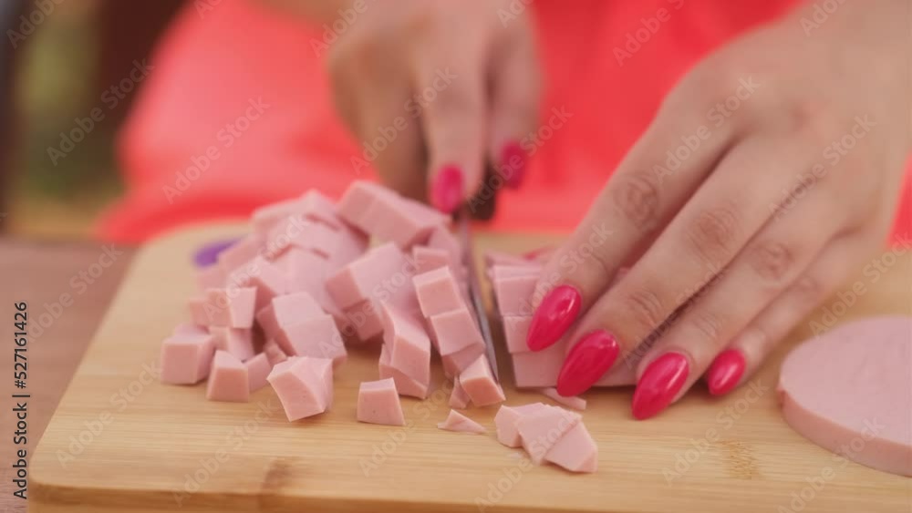 Unrecognizable lady cutting boiled sausage on wooden board outdoors. Woman's hands with bright manicure cutting sausage goods with knife on chopping board. Close up. Unrecognizable lady cutting boiled sausage on wooden board outdoors. Woman's hands with bright manicure cutting sausage goods with knife on chopping board. Close up.