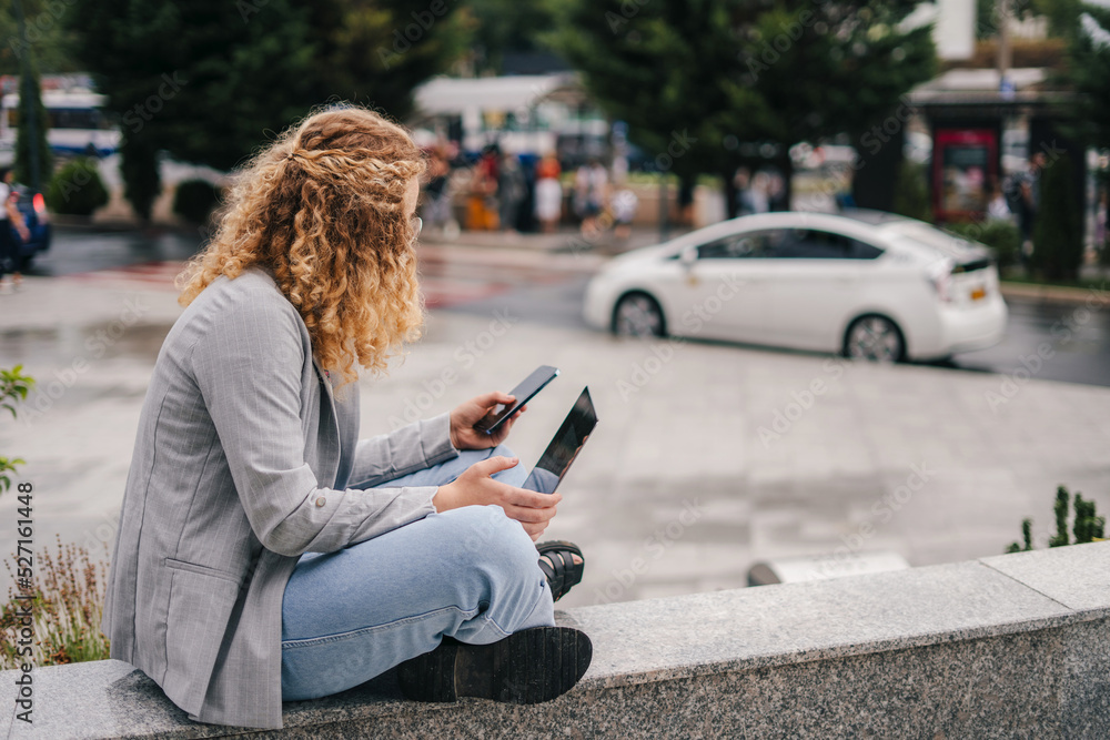 Side view blonde student woman sitting on the street and working on laptop, texting on the smart phone. Copy space. People urban lifestyle concept. Reading Side view blonde student woman sitting on the street and working on laptop, texting on the smart phone. Copy space. People urban lifestyle concept. Reading
