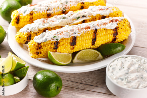 Elote on a white plate, grilled mexican street corn, roasted cobs are slathered in sour cream based sauce, seasoned with chili powder and sprinkled with cilantro.