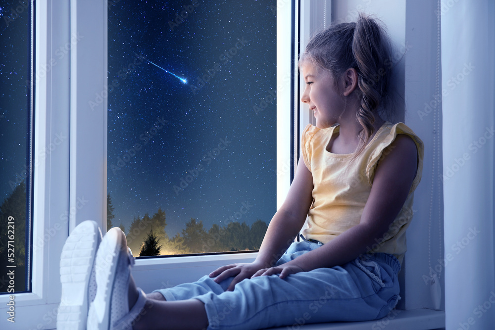 Cute little girl sitting near window and looking at shooting star in ...