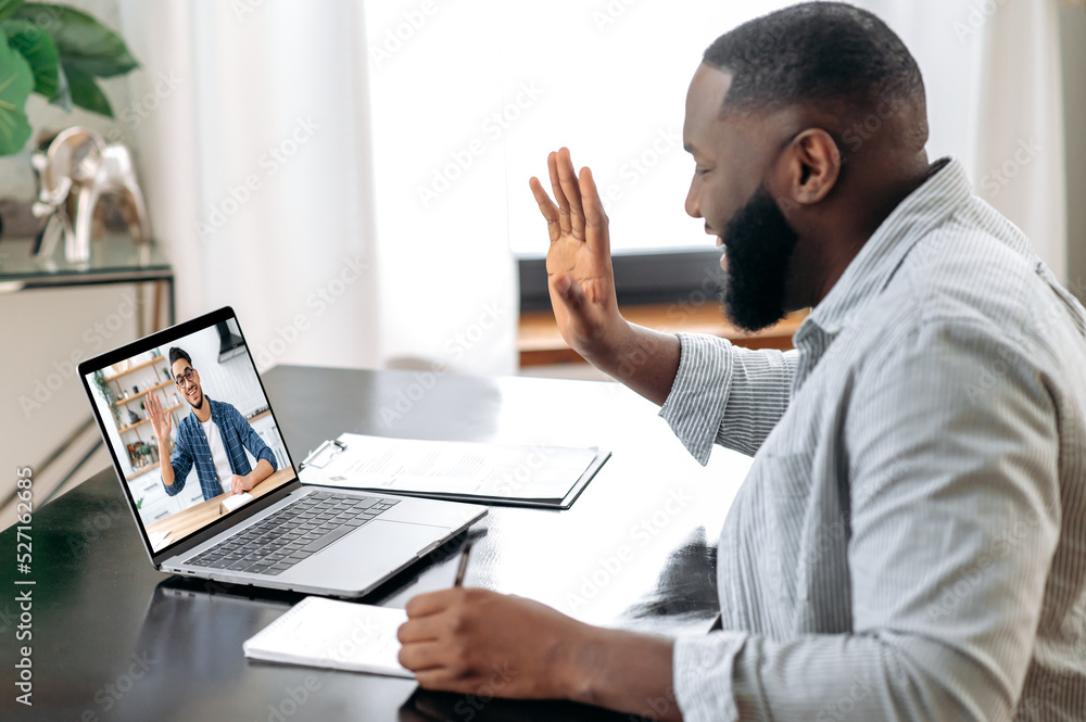 Online video meeting. African American young successful man talking on video conference with his colleague, friend or mentor while sitting at the desk in the office, talking about strategy,takes notes - obrazy, fototapety, plakaty 