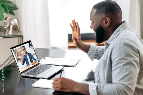 Online video meeting. African American young successful man talking on video conference with his colleague, friend or mentor while sitting at the desk in the office, talking about strategy,takes notes