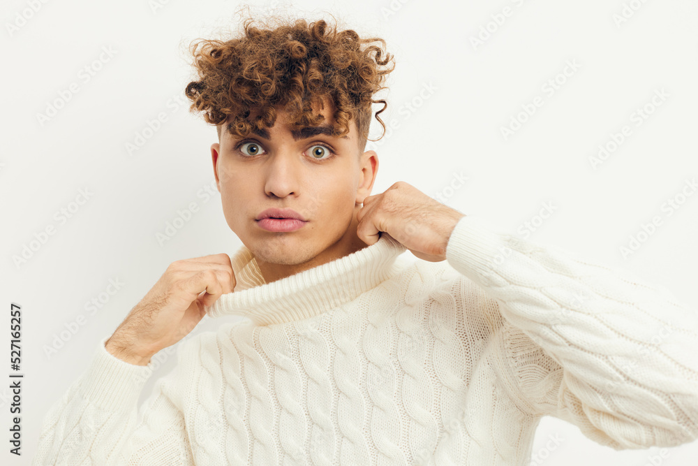 a close photo of a handsome, attractive man with curly hair, standing in a white knitted sweater on a light background, adjusting the neck of the sweater with his hands a close photo of a handsome, attractive man with curly hair, standing in a white knitted sweater on a light background, adjusting the neck of the sweater with his hands