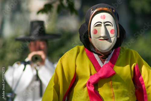 Person wearing a traditional South Korean mask while another man plays a tradtional instrument in the background © John