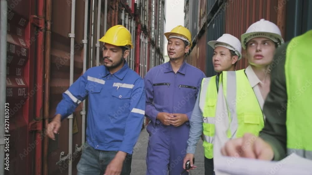 Group of multiracial workers in safety uniforms and hardhats walk and inspect shipping cargo with White male manager at nook of container stack, import and export goods logistic transport industry. Group of multiracial workers in safety uniforms and hardhats walk and inspect shipping cargo with White male manager at nook of container stack, import and export goods logistic transport industry.