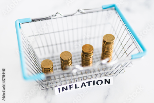 prices going up and rising inflation concept with text in front of shopping basket with growing stacks of coins inside of it