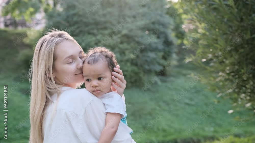 A Caucasian mother gently hugs her African child and holds him in her arms while walking in the park. Mixed multiracial family A Caucasian mother gently hugs her African child and holds him in her arms while walking in the park. Mixed multiracial family