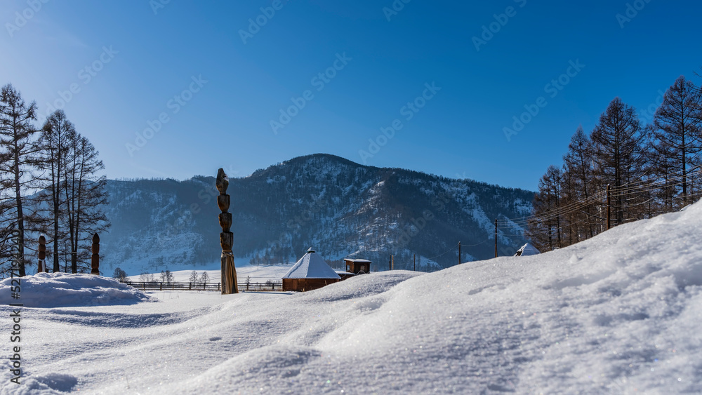 In the snow-covered valley there are traditional Altai houses - ail with a pyramidal roof. Pure white snow in the foreground. A mountain against a clear blue sky. Altai In the snow-covered valley there are traditional Altai houses - ail with a pyramidal roof. Pure white snow in the foreground. A mountain against a clear blue sky. Altai