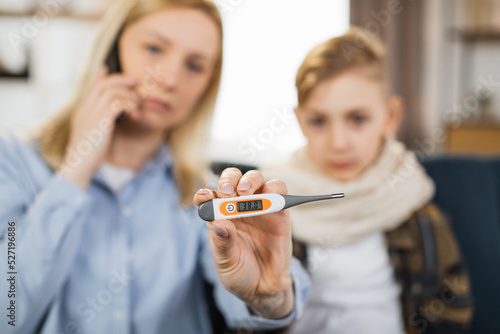 Mother showing to camera thermometer after measuring the temperature while using cell phone, calling to doctor. Mom checking temperature of her sick teen son with high fever sitting on sofa.