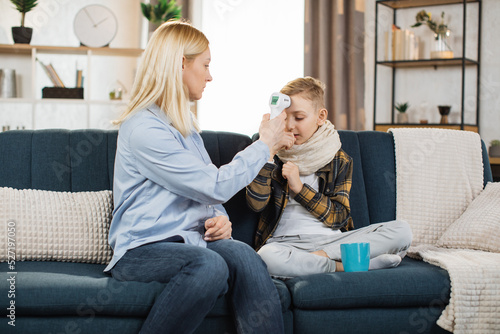 Middle aged worried caring blond woman, mother, sitting on sofa at home, hugging her sick sad teen son, measuring temperature using infrared wrist non-contact thermometer. Flu, cold, virus infection. photo