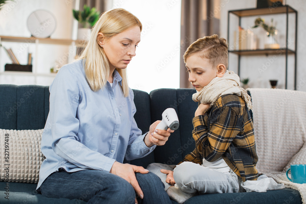 Mother taking care of her cute sick teen boy suffering from cold, checking temperature using infrared forehead thermometer gun, looking each other, sitting on the sofa. Mother taking care of her cute sick teen boy suffering from cold, checking temperature using infrared forehead thermometer gun, looking each other, sitting on the sofa.