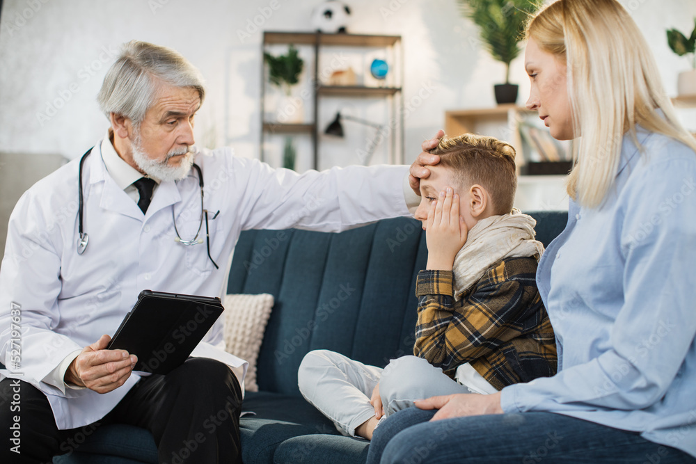Obraz premium Pleasant confident skilled mature man doctor in white coat, visiting patient cute sick preschool boy with his mother, at home, touches forehead to check temperature and giving recommendations.