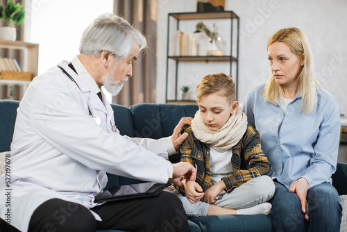 Front view of good-looking professional senior male caucasian doctor in medical coat, consulting his teen boy patient wihis mom, touching hand of child to check pulse, during home visit.