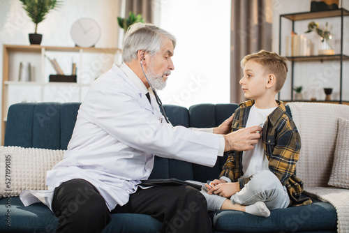 Preschool sick cute boy sitting on the couch while caring experienced nice mature male doctor listening his heartbeat during home visit, front view.