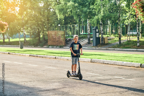 Schoolboy rides electric scooter on road in empty summer park. Child in black t-shirt and denim shorts rides on scooter along marked asphalt road © timltv