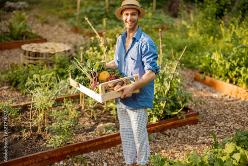 Portrait of young male farmer holds box of freshly picked vegetables while standing at local farmland. Concept of organic food grown and sustanable lifestyle
