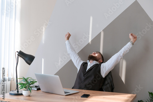 Portrait smiling man with beard, working in office on some project, he sits at table with a laptop, enjoys his success with his hands raised. Caucasian successful business guy