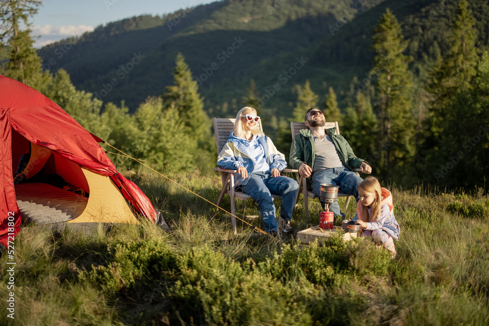 Young caucasian couple with little girl sit relaxed on chairs while traveling with tent in the mountains. Concept of happy family vacation on nature