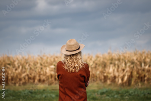 Woman with red coat and cowboy hat looks at cloudy sky over corn field