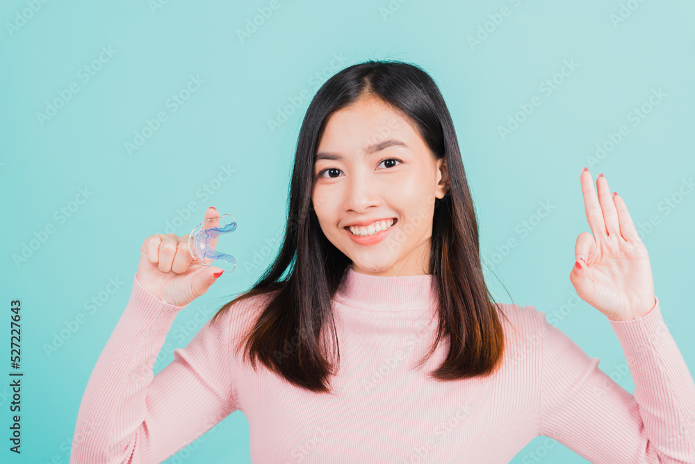 Portrait Asian beautiful young woman smiling holding silicone orthodontic retainers for teeth and show finger brackets saying OK sign, isolated on blue background, Dental hygiene healthy care concept Portrait Asian beautiful young woman smiling holding silicone orthodontic retainers for teeth and show finger brackets saying OK sign, isolated on blue background, Dental hygiene healthy care concept