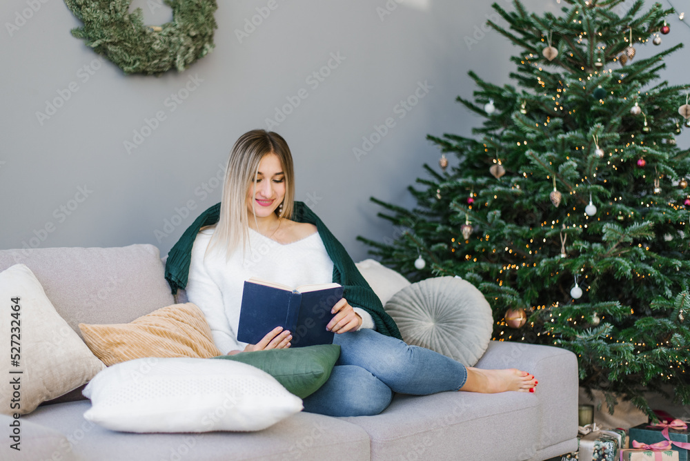 Beautiful woman is reading a book, resting on pillows with a blanket at the Christmas tree at home. Beautiful woman is reading a book, resting on pillows with a blanket at the Christmas tree at home.