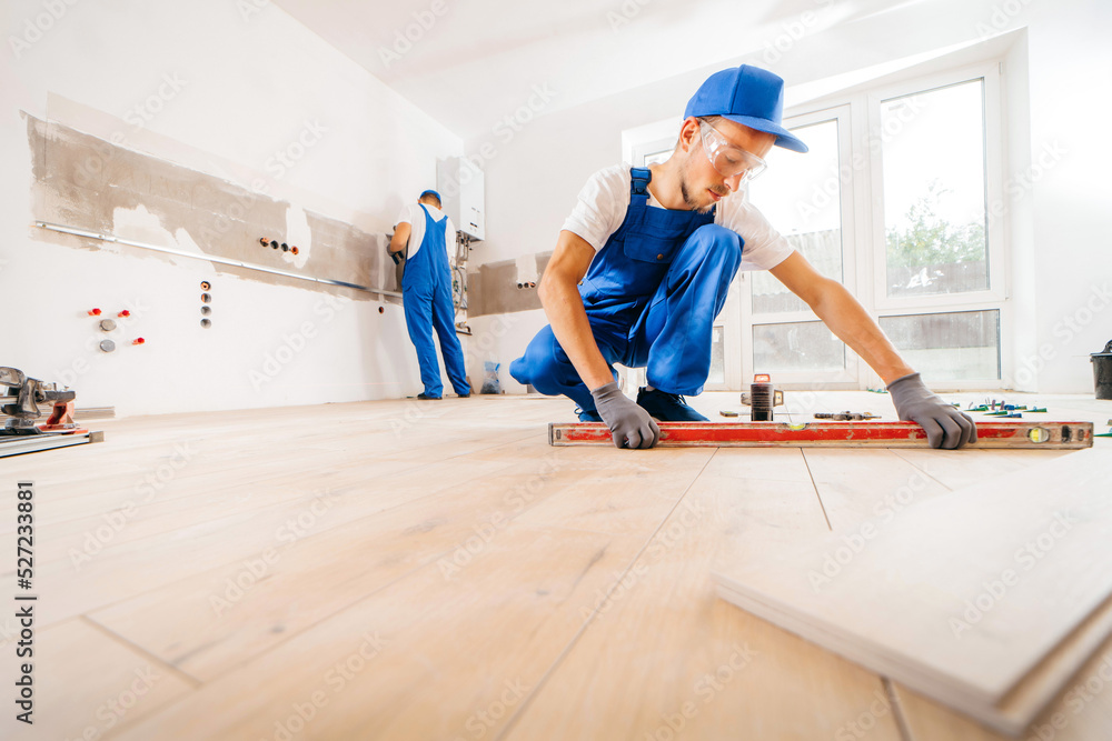 Two workers in a special uniform laying tiles with tile leveling system and laser level on the floor in a new house