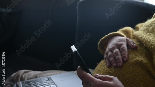 Pregnant woman sitting in the sofa using smart phone while stroking her belly. Concept of expecting woman working from home using laptop and mobile phone. Footage made in Sweden.