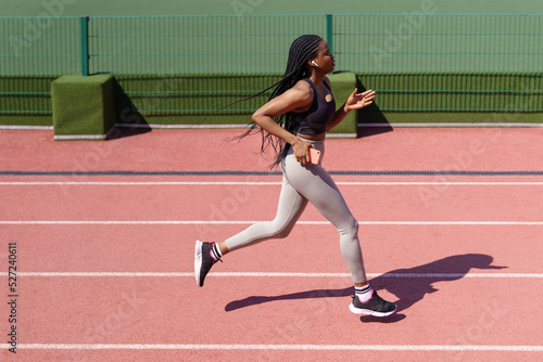 African American female enjoys running every day on red track listening to music in headphones on hot summer day. Young woman tries to keep healthy lifestyle at sports ground of residential complex