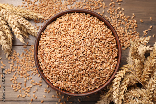 Wheat grains in bowl and spikes on wooden table, flat lay