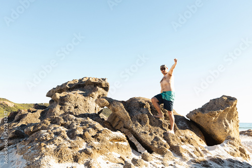 a father wearing board shorts and no shirt standing on top of rocks at a beach