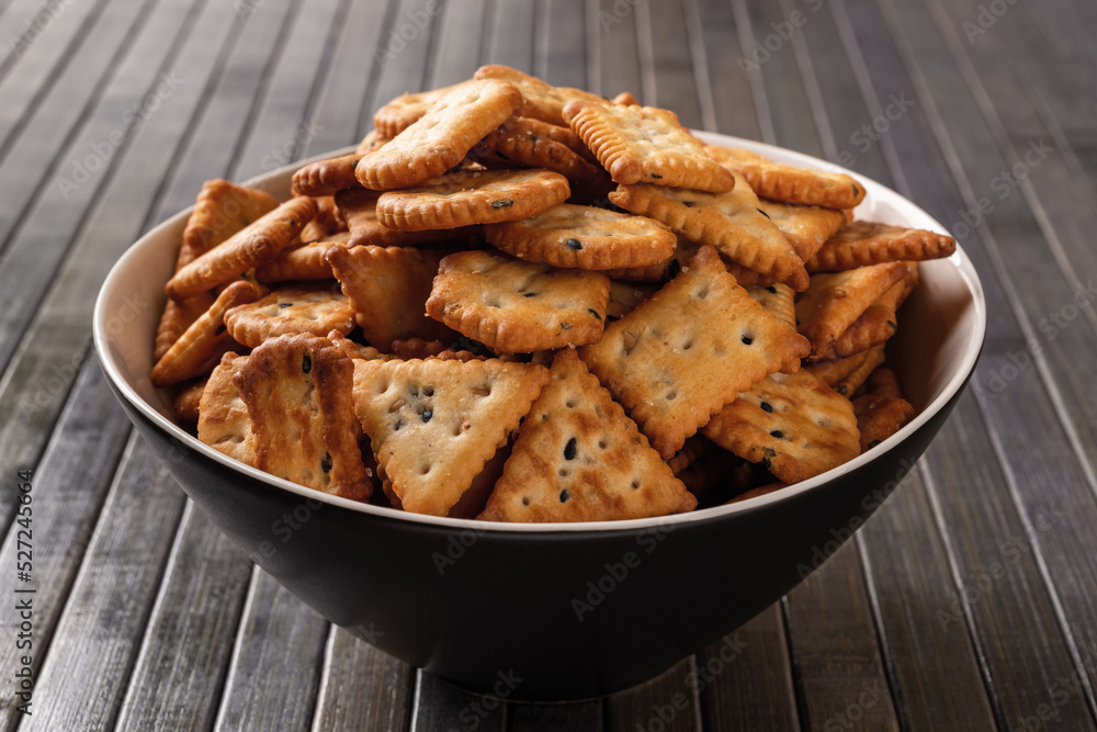 Heap of saltine crackers in a bowl over dark wood plank background. Black deep plate full of salty crackers with black and white sesame seeds. Baked crunchy snack. Saltines concept. Closeup. Heap of saltine crackers in a bowl over dark wood plank background. Black deep plate full of salty crackers with black and white sesame seeds. Baked crunchy snack. Saltines concept. Closeup.