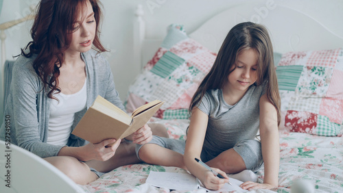 Young mother helps her little cute daughter with homework for elementary school. Loving mom reading a book and girl writing notes in copybook while sitting together on bed in cozy bedroom at home