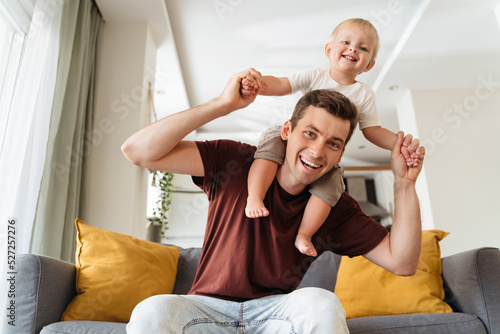 Happy young handsome dad carrying his baby boy on shoulders sitting on sofa posing in front of camera in living-room, both smiling and having fun while mom resting. Father having fun with his toddler