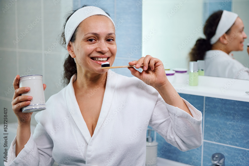 Happy Latin American woman in white bathrobe, brushing teeth with plastic free toothbrush and whitening toothpaste smiling beautiful toothy smile while looking at camera. Oral hygiene and dental care Happy Latin American woman in white bathrobe, brushing teeth with plastic free toothbrush and whitening toothpaste smiling beautiful toothy smile while looking at camera. Oral hygiene and dental care