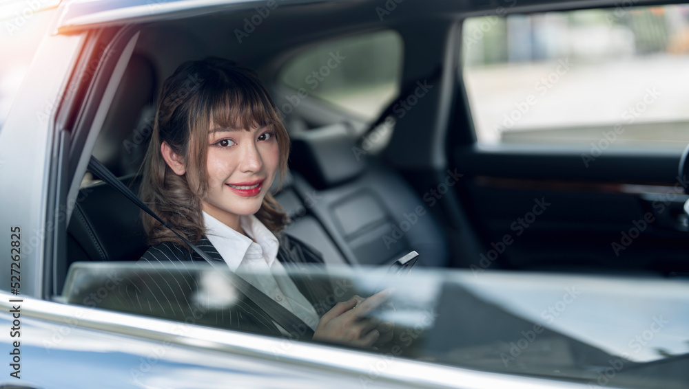 Beautiful asian woman using mobile phone while sitting in the back seat of her car. Smiling and looking at camera.