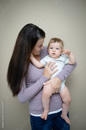 pretty young mother with dark hair, purple top is holding her 7 months old baby and standing in front of brown background and is happy and full of love