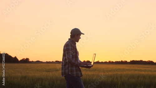 Farmer with a laptop computer in front of a sunset agricultural landscape. Man in a countryside field. Country life, food production, farming and technology concept.