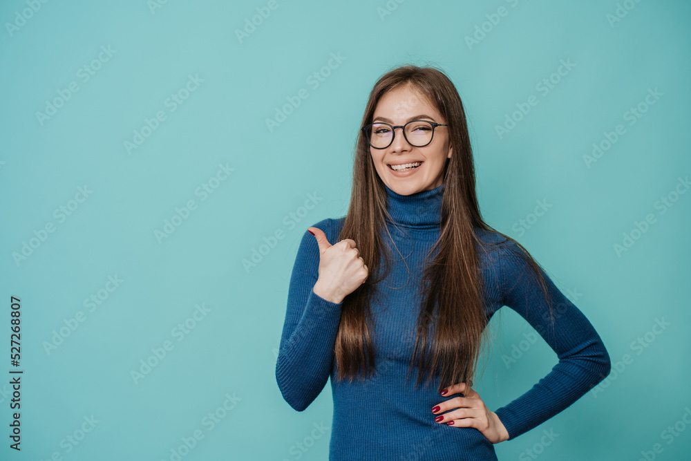 Attractive Italian young caucasian woman in blue sweater and glasses smiling wide, showing thumb up gesture over studio turquoise background with empty space for ad. Mockup, shopping, sales, discount. Attractive Italian young caucasian woman in blue sweater and glasses smiling wide, showing thumb up gesture over studio turquoise background with empty space for ad. Mockup, shopping, sales, discount.
