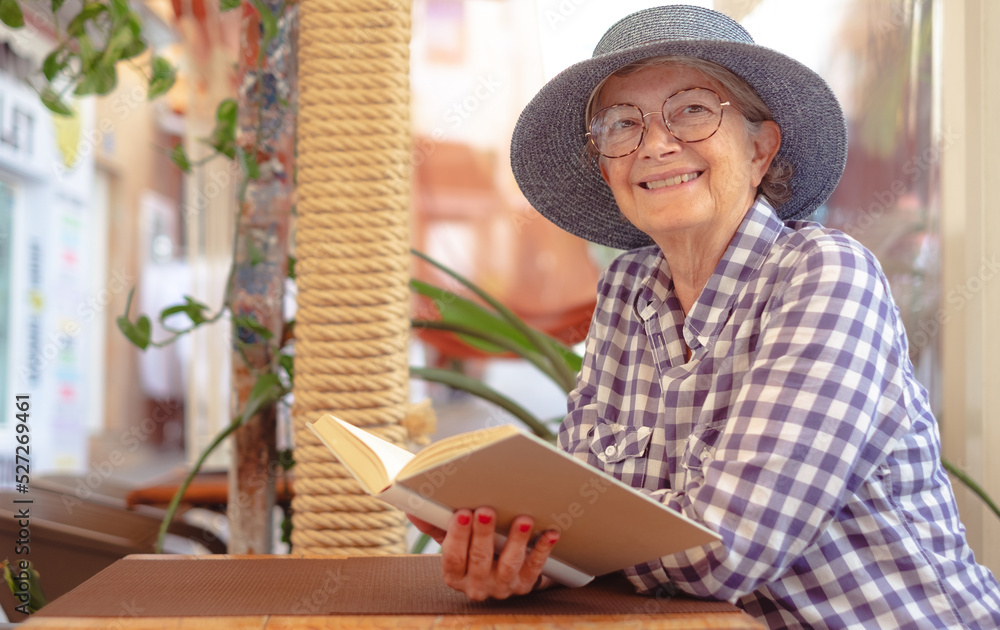 Portrait of beautiful senior woman with hat sitting at cafe table reading a book, studying - caucasian lady enjoying free time and retirement