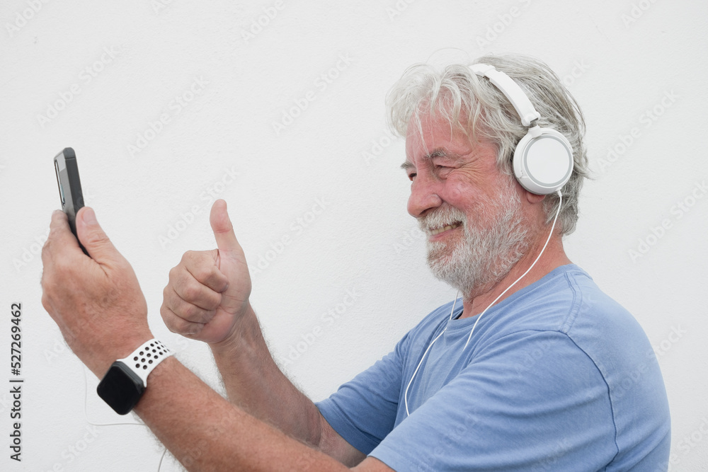 Smiling senior man wearing headphones in video call with phone and thumb up, copy space - attractive bearded joyful senior male enjoying tech and social Smiling senior man wearing headphones in video call with phone and thumb up, copy space - attractive bearded joyful senior male enjoying tech and social