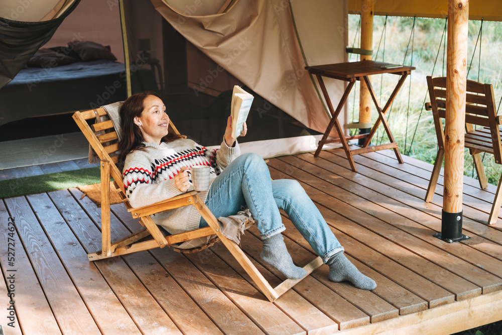 Brunette woman in nordic sweater reading book and relaxing in glamping in nature