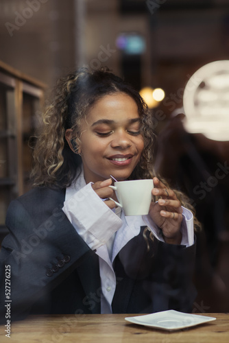 pleased african american woman in wireless earphone listening music and holding cup of coffee in cafe.